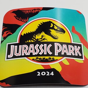 May include: A yellow, green, and red Jurassic Park logo with a black dinosaur silhouette and the text "Jurassic Park 2024".