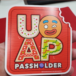 May include: A red and white gingerbread cookie sticker with the letters "UAP" and a gingerbread man face. The sticker says "Passholder" and includes a copyright notice.