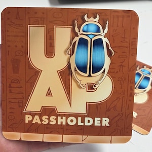 May include: A square card with a brown background featuring Egyptian-style hieroglyphs. The large letters "AP" are printed in a light tan color. A metallic-looking blue and gold scarab beetle is positioned over the letters. The word "PASSHOLDER" is printed in white at the bottom.