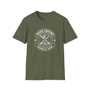 Pode incluir: Uma t-shirt de manga curta verde-oliva com um gráfico circular branco no peito. O design mostra duas ferramentas de escultura em madeira cruzadas sobre um padrão de grão de madeira, com o texto "WOOD CARVING" e "SHAPED BY HAND" na borda.