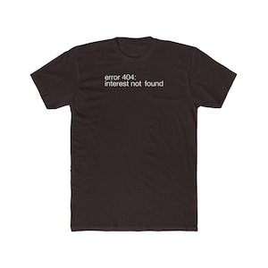 Op de afbeelding: Een donkerbruin t-shirt met korte mouwen en de witte tekst "error 404: interest not found" op de borst gedrukt. Het kledingstuk heeft een ronde hals en een standaard pasvorm, met een eenvoudig grafisch ontwerp in het midden op de voorzijde.