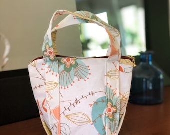 May include: A small white tote bag with a floral pattern in shades of orange, green, and blue. The bag has two handles and a zipper closure.