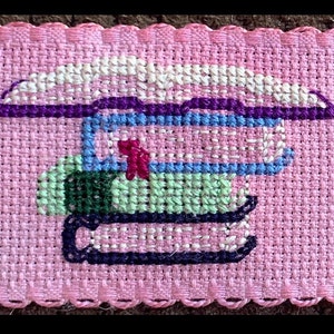 May include: A pink fabric with a cross-stitch design of a stack of books. The books are in shades of green, blue, purple, and white.