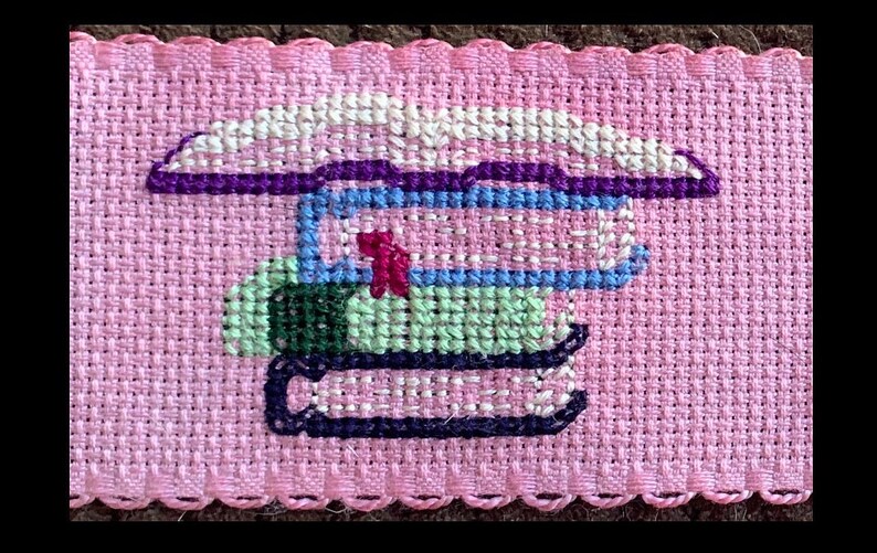 May include: A pink fabric with a cross-stitch design of a stack of books. The books are in shades of green, blue, purple, and white.