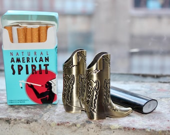 May include: A pack of Natural American Spirit cigarettes with a blue and white design featuring a Native American figure. Two gold-colored cowboy boot-shaped cigarette lighters are in the foreground. A black lighter is also visible.