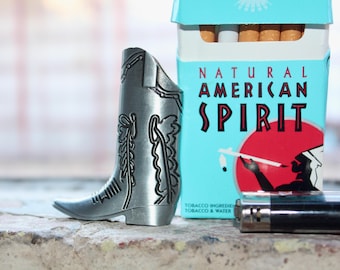 May include: A silver-colored cowboy boot-shaped lighter stands next to a Natural American Spirit cigarette pack. The pack is light blue with red and black text and a Native American graphic. A silver lighter is also visible.