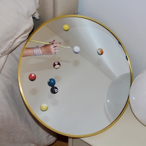 May include: A round mirror with a gold frame reflecting a pool table with colourful billiard balls and a cue stick. The mirror is on a white surface.