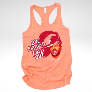 May include: A peach-colored racerback tank top featuring a graphic of a person wearing a pirate hat with the text "BUC AROUND AND FIND OUT" in red lettering, along with a signature below the face and the number 6 on the chin area of the illustration.