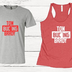 May include: A heather gray t-shirt and a red tank top. Both shirts feature the text "TOM BUC'ING BRADY" in a red and white design. The tank top has a racerback style.
