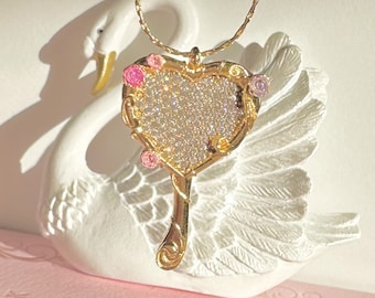May include: A gold heart-shaped mirror necklace with a sparkly surface and pink and white accents. The necklace is hanging from a white swan figurine.