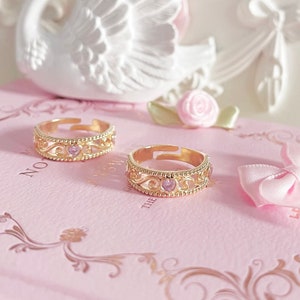 Pode incluir: Duas alianças de ouro com delicados desenhos em filigrana e pedras preciosas cor-de-rosa. As alianças estão sobre um livro rosa com as palavras "No" e "The" visíveis.