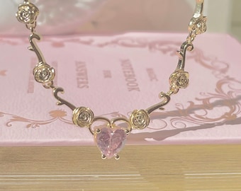 May include: A gold necklace with a pink heart-shaped pendant and rose accents. The necklace is on a pink book with the word "notebook" printed on the cover.