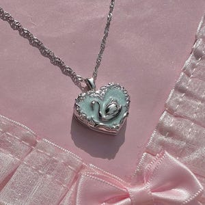 May include: A silver-toned heart-shaped locket pendant on a twisted chain. The locket features an ornate silver border surrounding a pale mint green enamel center with a raised silver swan motif in the middle, resting against a pink fabric background.