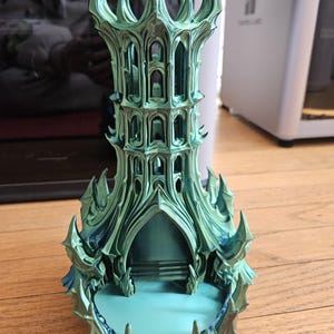 May include: A teal and blue decorative tower with a gothic architectural design. The tower features intricate details, including arched windows, spires, and a base with a textured surface. The object is made of a 3D printed material.