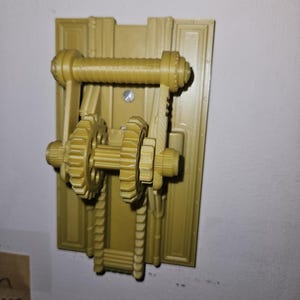 Könnte beinhalten: Ein goldfarbenes, 3D-gedrucktes Wanddekorationsstück mit einem mechanischen Zahnraddesign. Das Stück umfasst eine rechteckige Rückplatte, einen horizontalen Zylinder und zwei markante Zahnräder, die eine Steampunk-Ästhetik erzeugen.