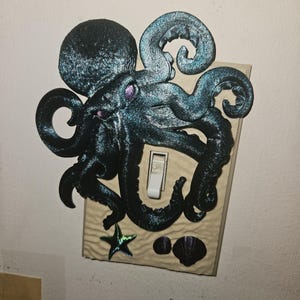 May include: Decorative light switch cover featuring a detailed, dark teal octopus design. The octopus wraps around the switch, with a starfish and seashell accents. The switch is in the "off" position.