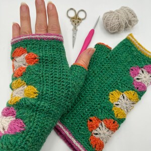 May include: A pair of green fingerless gloves with a crocheted flower design in pink, orange, yellow, and white. The gloves have a pink and white trim.