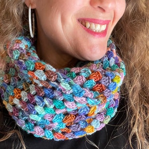 May include: A colorful crocheted neck warmer in shades of blue, green, purple, orange, and brown. The neck warmer is made with a textured stitch and is worn by a person with long, curly hair.