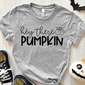 Puede incluir: Una camiseta gris con el texto "hey there pumpkin" en letras negras y un peque&ntilde;o gr&aacute;fico de calabaza. La camiseta est&aacute; colocada sobre una superficie de madera con un par de zapatillas negras y una calabaza blanca con una cara negra.