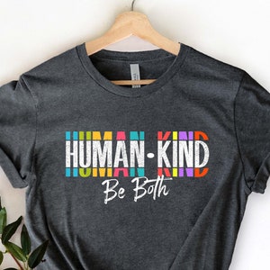 May include: A dark heather grey t-shirt with the text "HUMAN-KIND Be Both" in colorful letters. The letters are stacked vertically and have a vintage, distressed look.