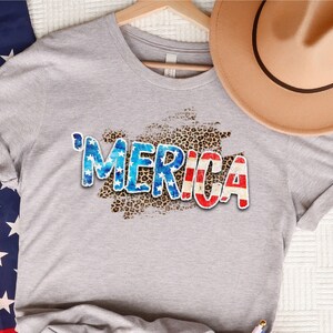 Op de afbeelding: Een grijs T-shirt met een verweerde "'MERICA"-print in rood, wit en blauw op een luipaardprint-achtergrond. Het shirt wordt gecombineerd met een spijkerbroekje en een rode bandana.