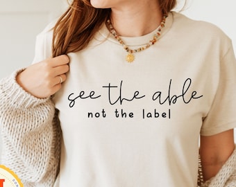 May include: A beige t-shirt with the text "see the able not the label" printed in black.