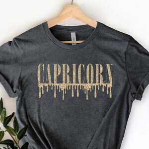 Pu&ograve; includere: Una maglietta grigio scuro con un design glitter oro che dice "CAPRICORNO" con gocce che cadono dalle lettere.