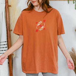 May include: Burnt orange t-shirt featuring an embroidered design of two koi fish. The fish are white, orange, and black with red fins. The shirt has short sleeves and a crew neck.