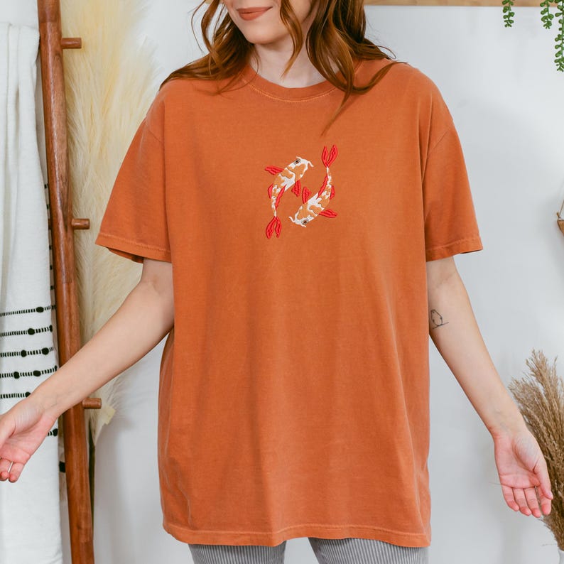 May include: Burnt orange t-shirt featuring an embroidered design of two koi fish. The fish are white, orange, and black with red fins. The shirt has short sleeves and a crew neck.