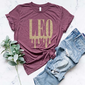 May include: A maroon t-shirt with a gold glitter design that says "LEO" with drips coming from the letters.