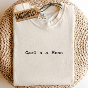 May include: Cream-colored t-shirt with the text "Carl's a Mess" in black. A brown tag that says "EMBROIDERY" is attached to the shirt. The shirt is folded on a woven, light brown surface.