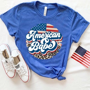 May include: A royal blue t-shirt with a white, red, and blue American flag design and the text "American Babe" in a retro font. The design is surrounded by a leopard print pattern.