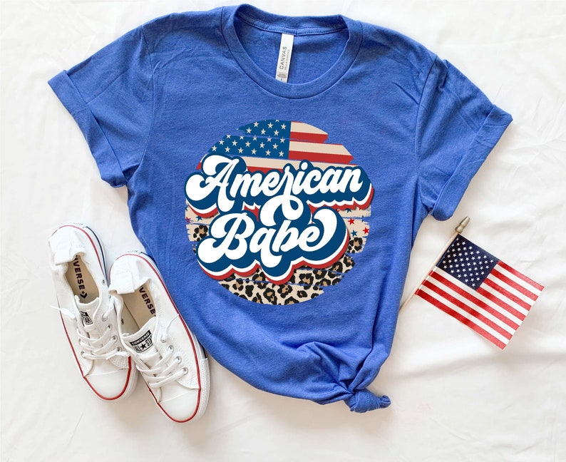 May include: A royal blue t-shirt with a white, red, and blue American flag design and the text "American Babe" in a retro font. The design is surrounded by a leopard print pattern.