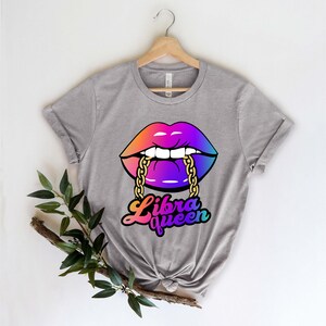 K&ouml;nnte beinhalten: Graues T-Shirt mit einer bunten Grafik von einem Paar Lippen, die eine goldene Kette bei&szlig;en. Der Text "Libra Queen" ist unter den Lippen gedruckt.