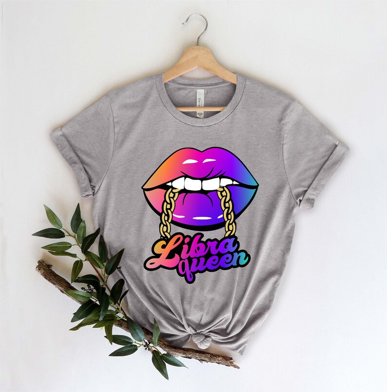 K&ouml;nnte beinhalten: Graues T-Shirt mit einer bunten Grafik von einem Paar Lippen, die eine goldene Kette bei&szlig;en. Der Text "Libra Queen" ist unter den Lippen gedruckt.