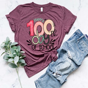 May include: A maroon t-shirt with the text "Happy 100th day of school" in colorful, handwritten lettering.