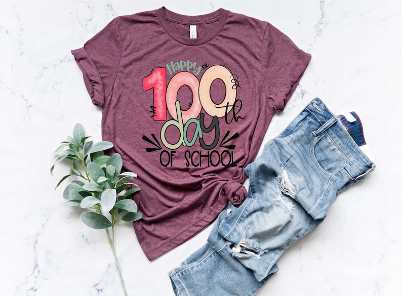 May include: A maroon t-shirt with the text "Happy 100th day of school" in colorful, handwritten lettering.