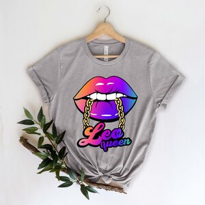 Pu&ograve; includere: Una maglietta grigia con una grafica arcobaleno di un paio di labbra con una catena dorata in bocca. Il testo "Leo queen" &egrave; sotto le labbra.