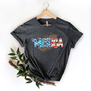Op de afbeelding: Een donkergrijs T-shirt met een verweerd Amerikaans vlagontwerp met de tekst "'MERICA" in witte letters op een luipaardprint achtergrond.