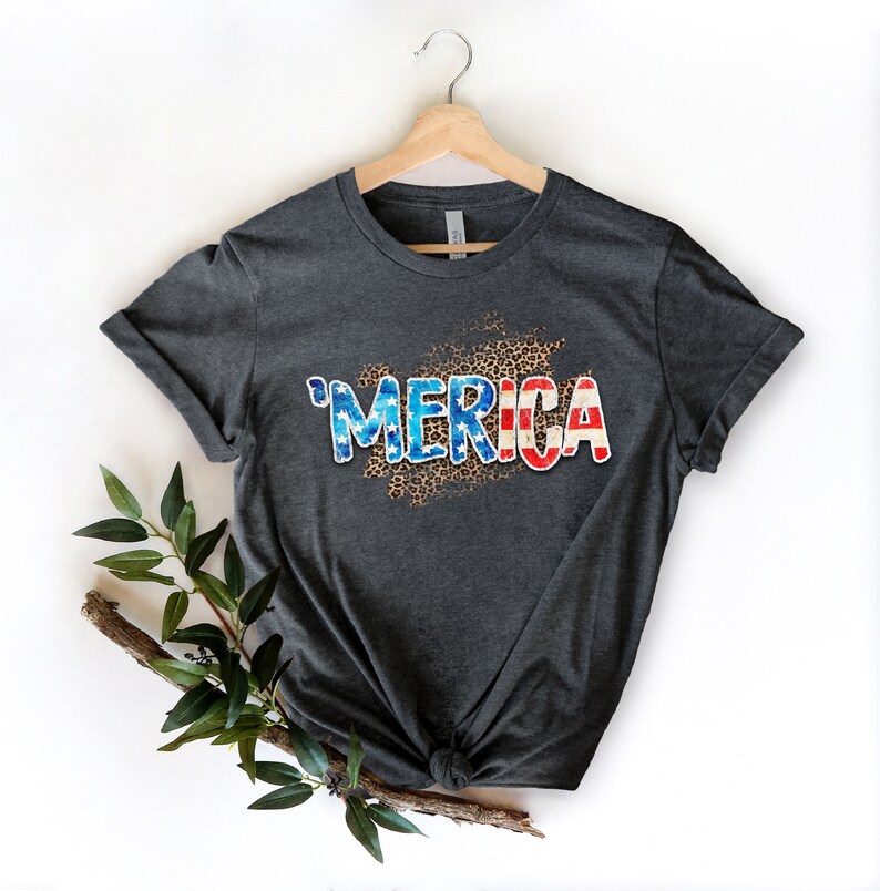 Op de afbeelding: Een donkergrijs T-shirt met een verweerd Amerikaans vlagontwerp met de tekst "'MERICA" in witte letters op een luipaardprint achtergrond.
