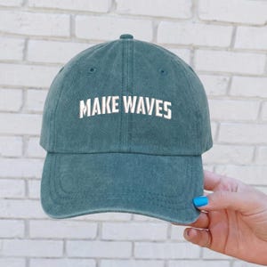 May include: A teal baseball cap with the phrase "MAKE WAVES" in white block letters across the front. The cap has a curved brim and a classic six-panel design. The background is a white brick wall.
