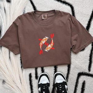 May include: A brown t-shirt featuring an embroidered design of two koi fish in orange, white, and red. The shirt is short-sleeved and has a crew neck. Black and white sneakers are visible at the bottom of the image.