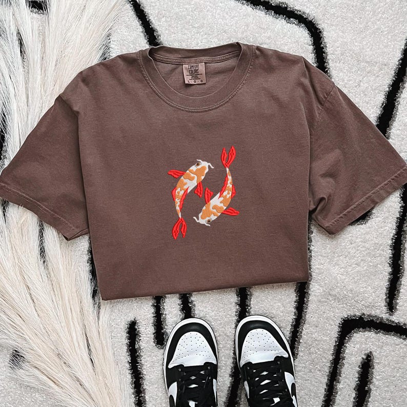 May include: A brown t-shirt featuring an embroidered design of two koi fish in orange, white, and red. The shirt is short-sleeved and has a crew neck. Black and white sneakers are visible at the bottom of the image.