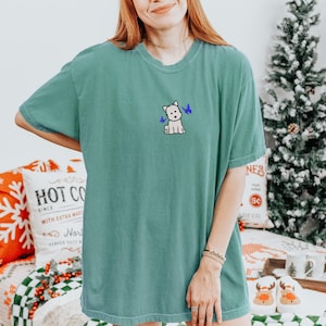 May include: A teal-colored t-shirt featuring a small embroidered design of a white dog with blue butterflies. The shirt has short sleeves and a crew neck. The image also shows a Christmas tree and a pillow with the text "HOT CO" on it.