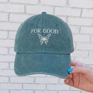 May include: A teal baseball cap with the words "FOR GOOD" in white script above a white butterfly graphic. The cap has a curved brim and a fabric strap closure.