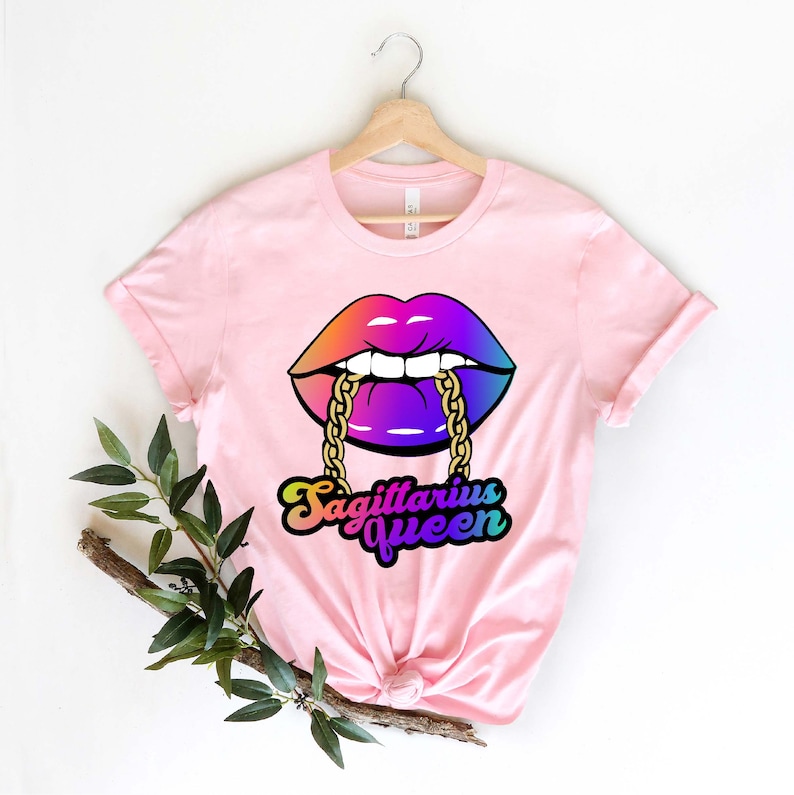 May include: Pink t-shirt with a rainbow-colored lips design and a gold chain. The text "Sagittarius Queen" is written in a rainbow-colored font below the lips.