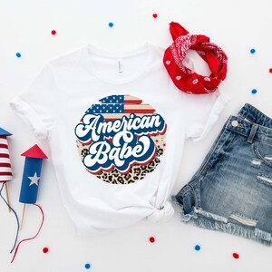 May include: White t-shirt with a red, white, and blue American flag design and the text "American Babe" in a retro font. The design is surrounded by a leopard print pattern.