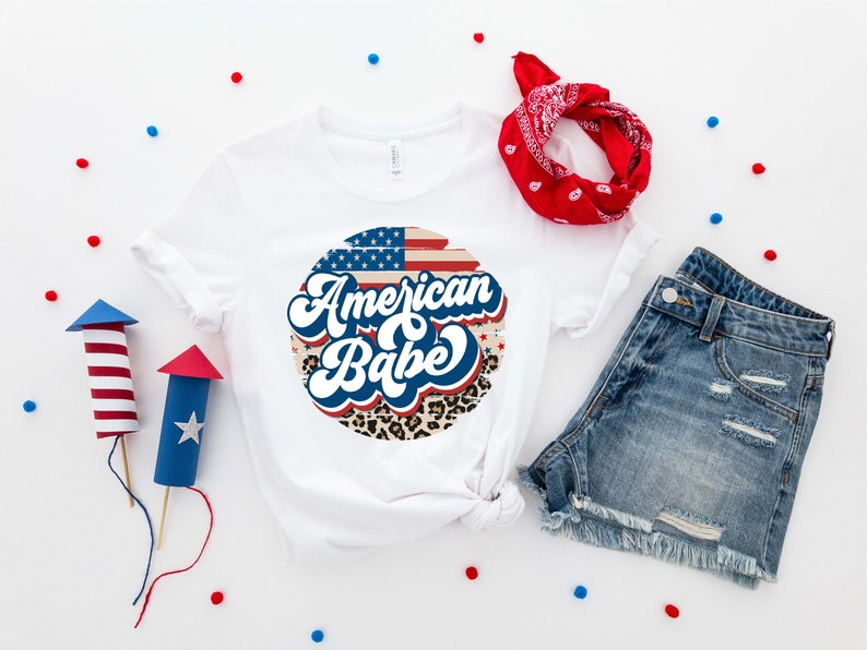 May include: White t-shirt with a red, white, and blue American flag design and the text "American Babe" in a retro font. The design is surrounded by a leopard print pattern.