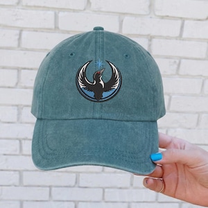 May include: Teal baseball cap with a circular embroidered design featuring a black bird with white wings and a red eye, set against a blue and white background. The cap has a curved brim and a fabric strap closure.