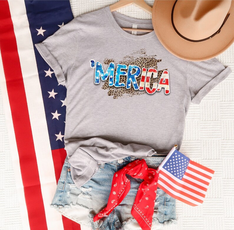 Op de afbeelding: Een grijs T-shirt met een verweerde "'MERICA"-print in rood, wit en blauw op een luipaardprint-achtergrond. Het shirt wordt gecombineerd met een spijkerbroekje en een rode bandana.